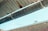 A metal gutter is attached to the edge of a corrugated roof, with water droplets dripping down. The background shows a blurred brick wall and a blue sky.