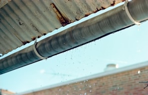 A metal gutter is attached to the edge of a corrugated roof, with water droplets dripping down. The background shows a blurred brick wall and a blue sky.