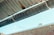 A metal gutter is attached to the edge of a corrugated roof, with water droplets dripping down. The background shows a blurred brick wall and a blue sky.