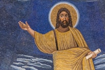 A mosaic artwork features a figure with long hair and a beard, wearing a golden robe, and surrounded by a halo. The figure holds a scroll in one hand while the other hand is raised in a gesture. The background consists of shades of blue, with cloud-like formations at the bottom.