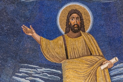 A mosaic artwork features a figure with long hair and a beard, wearing a golden robe, and surrounded by a halo. The figure holds a scroll in one hand while the other hand is raised in a gesture. The background consists of shades of blue, with cloud-like formations at the bottom.