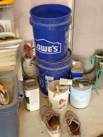A cluttered garage corner with two stacked blue Lowe's buckets, a worn pair of shoes on the floor, several paint cans with paintbrushes resting on top, and various tools and containers organized on shelves. There is a roll of paper towels and some garden equipment visible.
