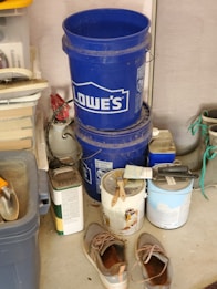 A cluttered garage corner with two stacked blue Lowe's buckets, a worn pair of shoes on the floor, several paint cans with paintbrushes resting on top, and various tools and containers organized on shelves. There is a roll of paper towels and some garden equipment visible.