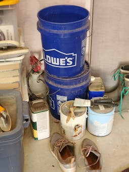 A cluttered garage corner with two stacked blue Lowe's buckets, a worn pair of shoes on the floor, several paint cans with paintbrushes resting on top, and various tools and containers organized on shelves. There is a roll of paper towels and some garden equipment visible.