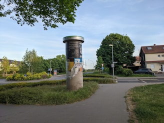 A cylindrical advertising column stands on a paved walkway surrounded by greenery and trees. The column is covered with torn and layered posters. In the background, there are residential buildings and a couple of street signs, including a yield sign. The scene is set in a suburban area with a clear blue sky overhead.