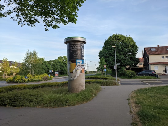 A cylindrical advertising column stands on a paved walkway surrounded by greenery and trees. The column is covered with torn and layered posters. In the background, there are residential buildings and a couple of street signs, including a yield sign. The scene is set in a suburban area with a clear blue sky overhead.