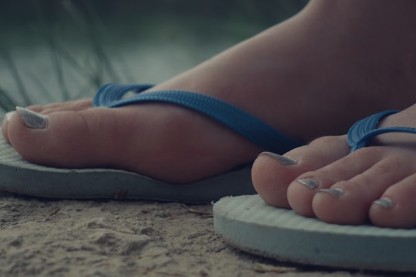 A close-up view of feet wearing light blue flip-flops, positioned on a sandy or earthy surface. The toenails are painted with a metallic color, and the background appears slightly blurred, suggesting outdoor scenery.