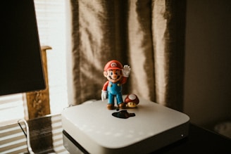 A Super Mario figurine stands on top of a sleek, silver Apple device, accompanied by a small mushroom figure. The scene is softly lit, with neutral tones from the curtain and background, creating a cozy, nostalgic atmosphere.