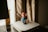 A Super Mario figurine stands on top of a sleek, silver Apple device, accompanied by a small mushroom figure. The scene is softly lit, with neutral tones from the curtain and background, creating a cozy, nostalgic atmosphere.