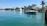 A serene coastal scene featuring a luxury motorboat carrying several people enjoying the sun. The boat is situated near lush green palm trees and a white, elegant waterfront house. Another yacht is visible in the background, docked near the shore.