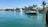 A serene coastal scene featuring a luxury motorboat carrying several people enjoying the sun. The boat is situated near lush green palm trees and a white, elegant waterfront house. Another yacht is visible in the background, docked near the shore.