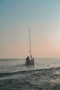 A small sailboat is on a calm sea under a vast, clear sky. Three individuals are aboard, working together as the boat gently navigates the water. The horizon is subtly visible, enveloped in a warm, soft light indicating either dawn or dusk.