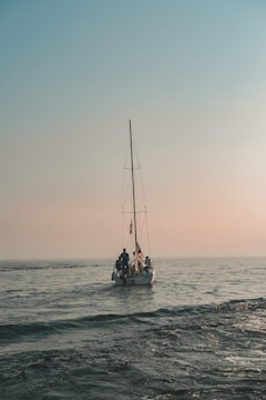 A small sailboat is on a calm sea under a vast, clear sky. Three individuals are aboard, working together as the boat gently navigates the water. The horizon is subtly visible, enveloped in a warm, soft light indicating either dawn or dusk.