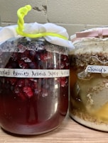 Two jars with cloth covers are filled with fermented contents. The left jar contains dark red liquid with visible berries and is labeled 'Fermented Berries Aronia Syrup.' The right jar contains a mixture with lighter colors and is labeled, having a creamy and grainy texture at the top.