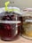 Two jars with cloth covers are filled with fermented contents. The left jar contains dark red liquid with visible berries and is labeled 'Fermented Berries Aronia Syrup.' The right jar contains a mixture with lighter colors and is labeled, having a creamy and grainy texture at the top.