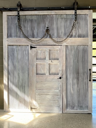 A large wooden sliding barn door with metal corrugated panels, set on a rail system. The door features visible chains and metal hardware, creating a rustic and industrial appearance. It has a smaller door within its structure.