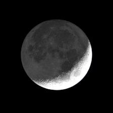 A detailed view of the moon with visible craters and surface texture, set against a pitch-black background. The lighting highlights the contours and topography, particularly emphasizing the right side of the lunar surface.