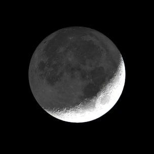 A detailed view of the moon with visible craters and surface texture, set against a pitch-black background. The lighting highlights the contours and topography, particularly emphasizing the right side of the lunar surface.