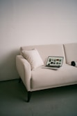 A minimalistic setting with a beige sofa placed against a plain wall. On the sofa, there is a laptop displaying images, along with a white cushion. A camera lens or similar object is placed beside the laptop.