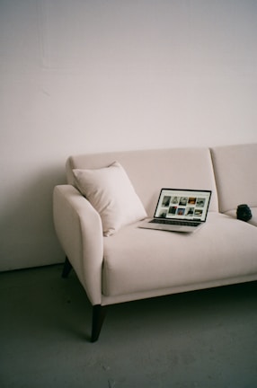 A minimalistic setting with a beige sofa placed against a plain wall. On the sofa, there is a laptop displaying images, along with a white cushion. A camera lens or similar object is placed beside the laptop.