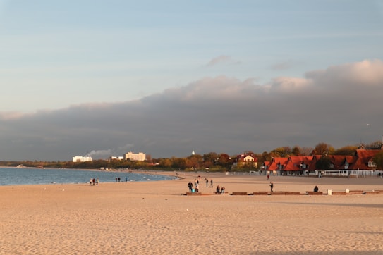 The image depicts a sandy beach with several people walking along the shore. In the background, there are buildings with red roofs surrounded by trees. The sky is mostly clear with some clouds, and the sun casts a warm light over the scene.