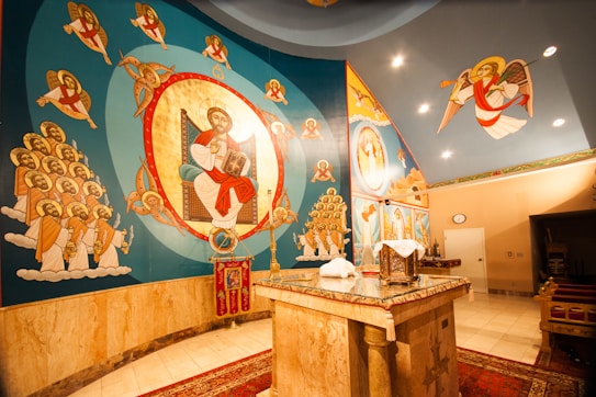 A vibrant interior of a religious space featuring elaborate wall paintings and murals depicting religious figures and scenes, with angels and a central figure holding a book. The walls are adorned with vivid colors and gold accents, creating an ornate and sacred atmosphere. The marble altar in the center is covered with cloth and religious artifacts.