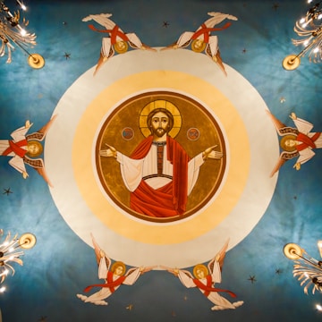 A circular religious mural on a ceiling features a central figure in red robes with open arms, surrounded by a halo. Four angels with red sashes and wings are symmetrically positioned around the figure, each floating in a different direction. The background is a gradient of blue, and there are decorative motifs and stars.