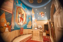 The image depicts an interior of a religious space with vibrant religious iconography on the walls. The artwork features various religious figures, including a central figure surrounded by an ornate design. The ceiling has additional religious symbolism and there is a ceremonial table in the center covered with cloths and some objects.