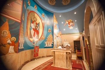 The image depicts an interior of a religious space with vibrant religious iconography on the walls. The artwork features various religious figures, including a central figure surrounded by an ornate design. The ceiling has additional religious symbolism and there is a ceremonial table in the center covered with cloths and some objects.