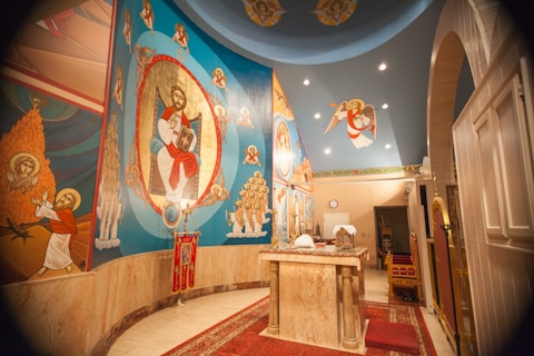 The image depicts an interior of a religious space with vibrant religious iconography on the walls. The artwork features various religious figures, including a central figure surrounded by an ornate design. The ceiling has additional religious symbolism and there is a ceremonial table in the center covered with cloths and some objects.