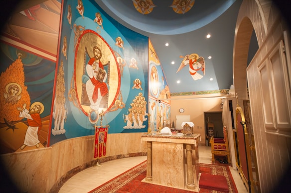 The image depicts an interior of a religious space with vibrant religious iconography on the walls. The artwork features various religious figures, including a central figure surrounded by an ornate design. The ceiling has additional religious symbolism and there is a ceremonial table in the center covered with cloths and some objects.
