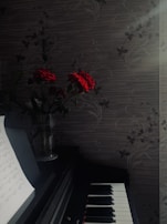 A darkly lit setting features a piano with visible sheet music on a stand. Beside the piano, a vase holds red roses. The background consists of a patterned wall with a subtle floral design. Soft sunlight filters through, creating a serene atmosphere.