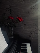 A darkly lit setting features a piano with visible sheet music on a stand. Beside the piano, a vase holds red roses. The background consists of a patterned wall with a subtle floral design. Soft sunlight filters through, creating a serene atmosphere.