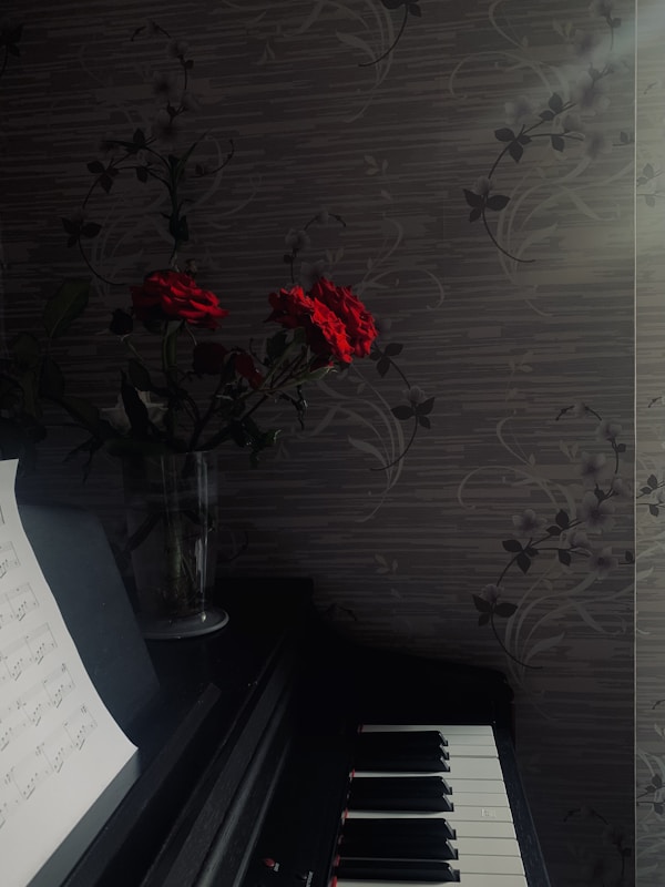 A darkly lit setting features a piano with visible sheet music on a stand. Beside the piano, a vase holds red roses. The background consists of a patterned wall with a subtle floral design. Soft sunlight filters through, creating a serene atmosphere.