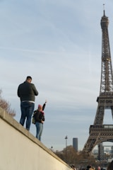 A person is standing on a platform with their back facing the camera, looking at a person in a pink hat and scarf who is gesturing enthusiastically. The Eiffel Tower occupies the right side of the image, towering against a blue sky with light clouds.