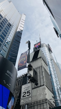 A cityscape featuring tall skyscrapers with glass facades looking upwards. There are advertisements displayed on digital billboards, including one for a musical. A speed limit sign is visible in the foreground.