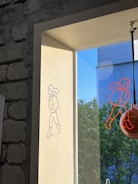 A wall with exposed bricks next to a window features a drawing of a simple cartoon figure walking, projected by light. The shadow of the figure appears on the wall, and another similar red figure is drawn on the window pane. The view outside reveals a building against a clear blue sky and green trees.