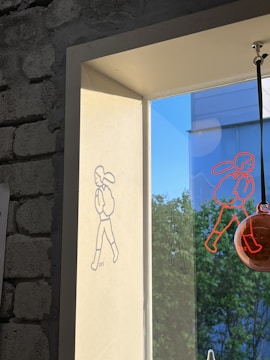 A wall with exposed bricks next to a window features a drawing of a simple cartoon figure walking, projected by light. The shadow of the figure appears on the wall, and another similar red figure is drawn on the window pane. The view outside reveals a building against a clear blue sky and green trees.