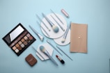 A flat lay of makeup products including a beige wallet, an eyeshadow palette, various makeup brushes with light blue handles, a compact, and a red lipstick all arranged on a light blue background.