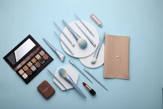 A flat lay of makeup products including a beige wallet, an eyeshadow palette, various makeup brushes with light blue handles, a compact, and a red lipstick all arranged on a light blue background.