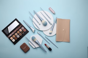 A flat lay of makeup products including a beige wallet, an eyeshadow palette, various makeup brushes with light blue handles, a compact, and a red lipstick all arranged on a light blue background.