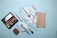 A flat lay of makeup products including a beige wallet, an eyeshadow palette, various makeup brushes with light blue handles, a compact, and a red lipstick all arranged on a light blue background.