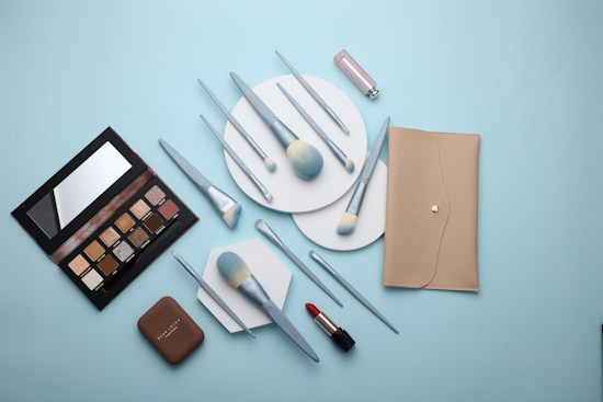 A flat lay of makeup products including a beige wallet, an eyeshadow palette, various makeup brushes with light blue handles, a compact, and a red lipstick all arranged on a light blue background.