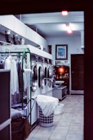 A laundry room with several industrial washing machines aligned against a wall. There's a laundry basket with clothes in front of the machines. A rack with hanging clothes is visible on the left side. The room is softly lit with fluorescent ceiling lights, and a framed picture hangs on the back wall next to some small decorative items on a shelf.