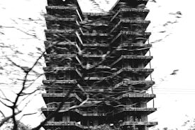 A high-rise building under construction with multiple floors visible, captured in black and white. The structure is shrouded partially by blurred tree branches, indicating movement or wind. The scene appears gritty and somewhat chaotic due to the stark contrast and dynamic composition.