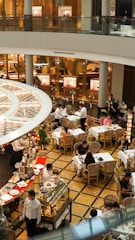 A bustling indoor cafe scene with multiple tables and patrons enjoying their meals. The area is decorated with elegant furniture and plants, and waitstaff are actively serving in the space. There is a display of pastries and desserts in the forefront, along with a set of commercial shop windows in the background.