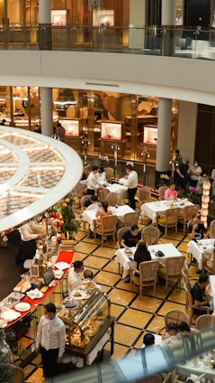 A bustling indoor cafe scene with multiple tables and patrons enjoying their meals. The area is decorated with elegant furniture and plants, and waitstaff are actively serving in the space. There is a display of pastries and desserts in the forefront, along with a set of commercial shop windows in the background.