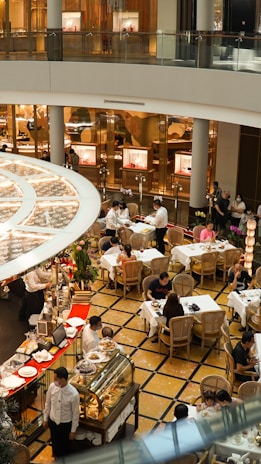 A bustling indoor cafe scene with multiple tables and patrons enjoying their meals. The area is decorated with elegant furniture and plants, and waitstaff are actively serving in the space. There is a display of pastries and desserts in the forefront, along with a set of commercial shop windows in the background.