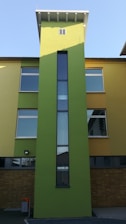 A modern building featuring a tall, narrow green tower with a vertical window at the center. The building has a facade with yellow and green sections and numerous rectangular windows. The base of the building is composed of brown bricks and there is a small trash bin visible in the foreground.