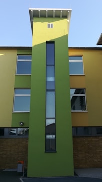A modern building featuring a tall, narrow green tower with a vertical window at the center. The building has a facade with yellow and green sections and numerous rectangular windows. The base of the building is composed of brown bricks and there is a small trash bin visible in the foreground.